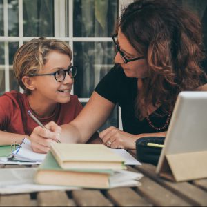 Young student doing homework at home with school books, newspaper and digital pad helped by his mother. Mum writing on the copybook teaching his son. Education, family lifestyle, homeschooling concept